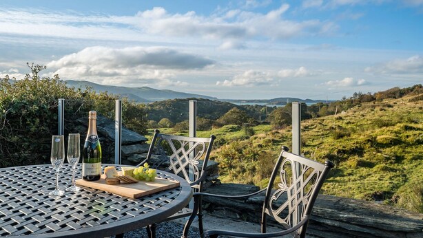 A table with a bottle of wine, glasses, cheese, and grapes set outdoors with a scenic view of hills and a clear sky.