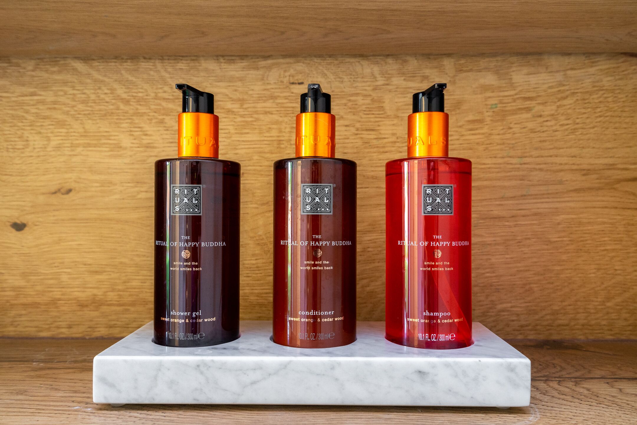 Three bottles of The Ritual of Happy Buddha products on a marble shelf.