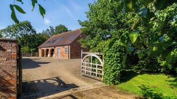 A brick building with a sloped roof and arched windows, surrounded by greenery and a cobblestone driveway.