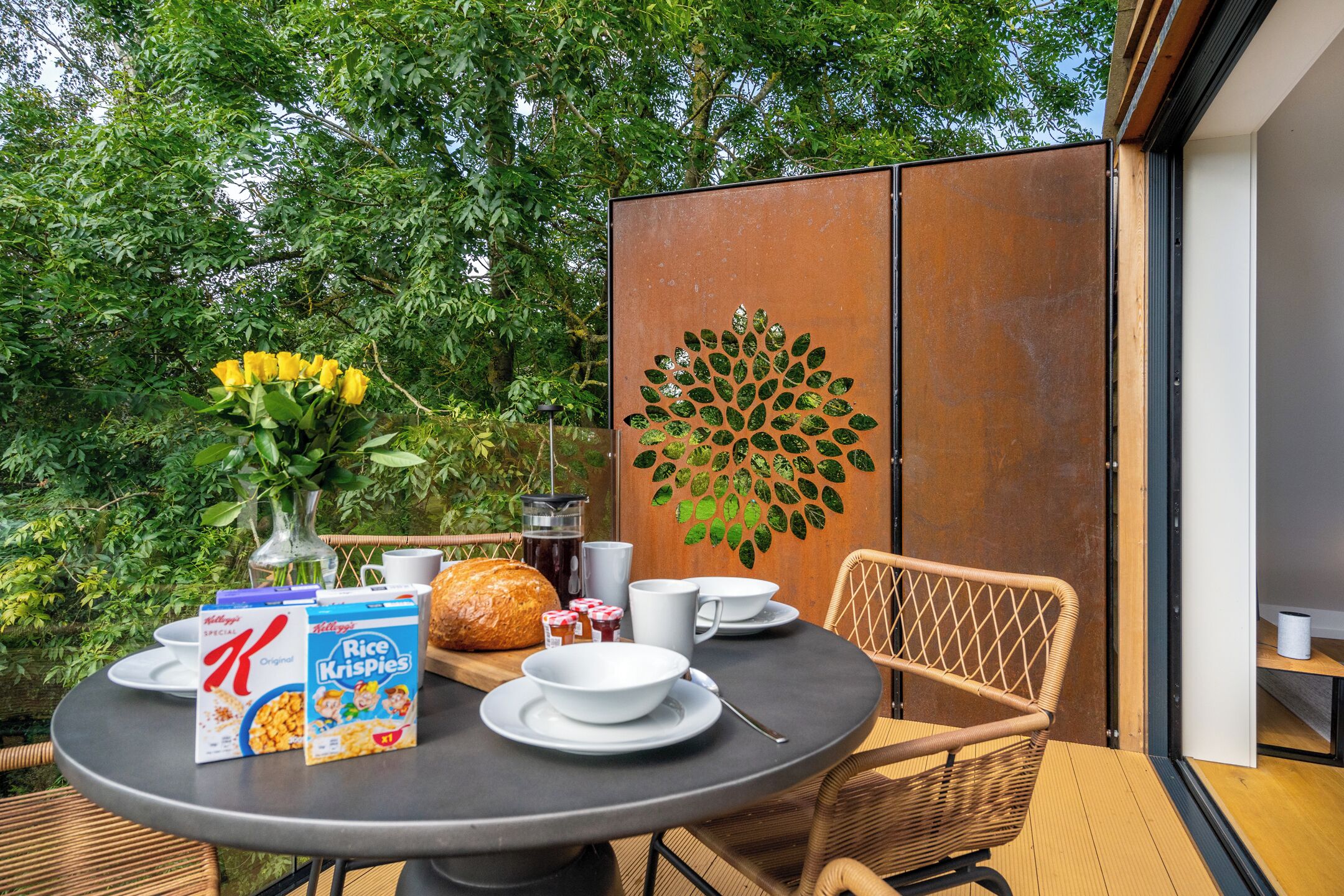 Une table ronde avec des aliments pour le petit-déjeuner, une chaise en osier et un mur décoratif orné d’un motif de feuilles.