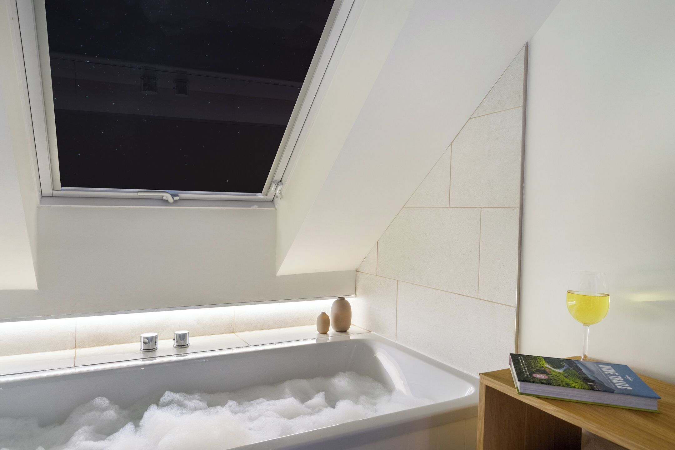 A modern bathroom with a bathtub filled with bubbles, a window above, and a glass of wine on a side table.