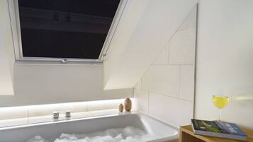 A modern bathroom with a bathtub filled with bubbles, a window above, and a glass of wine on a side table.
