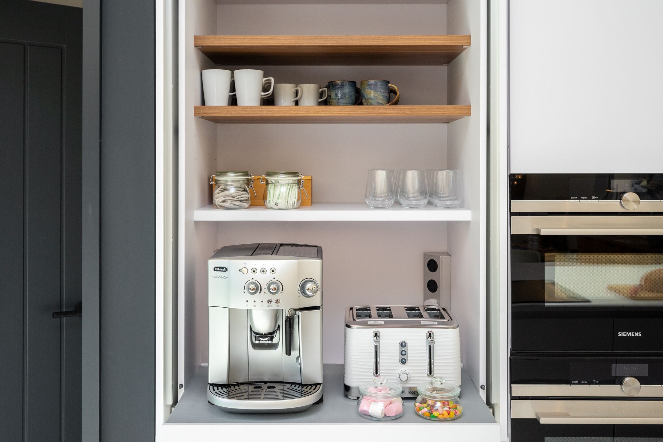 Una cocina con cafetera, tostadora y estantes con tazas y frascos.