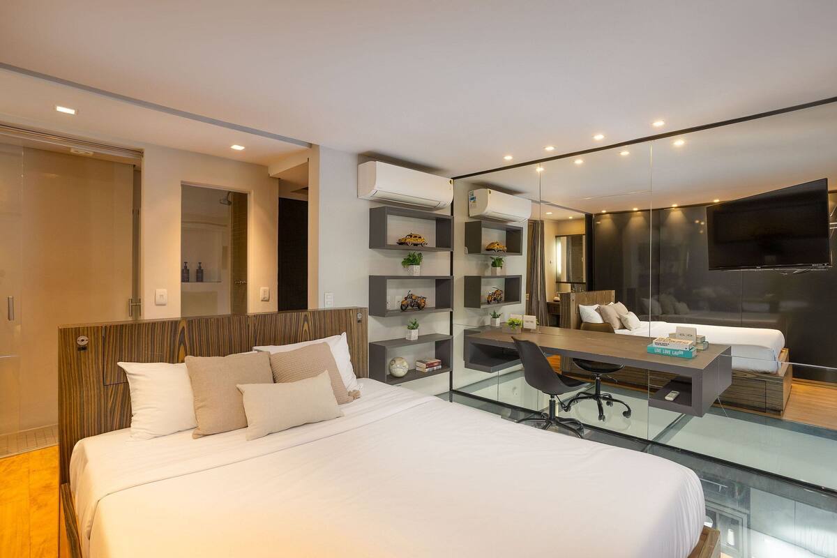 A modern hotel room with a large bed, a desk, and a glass partition.