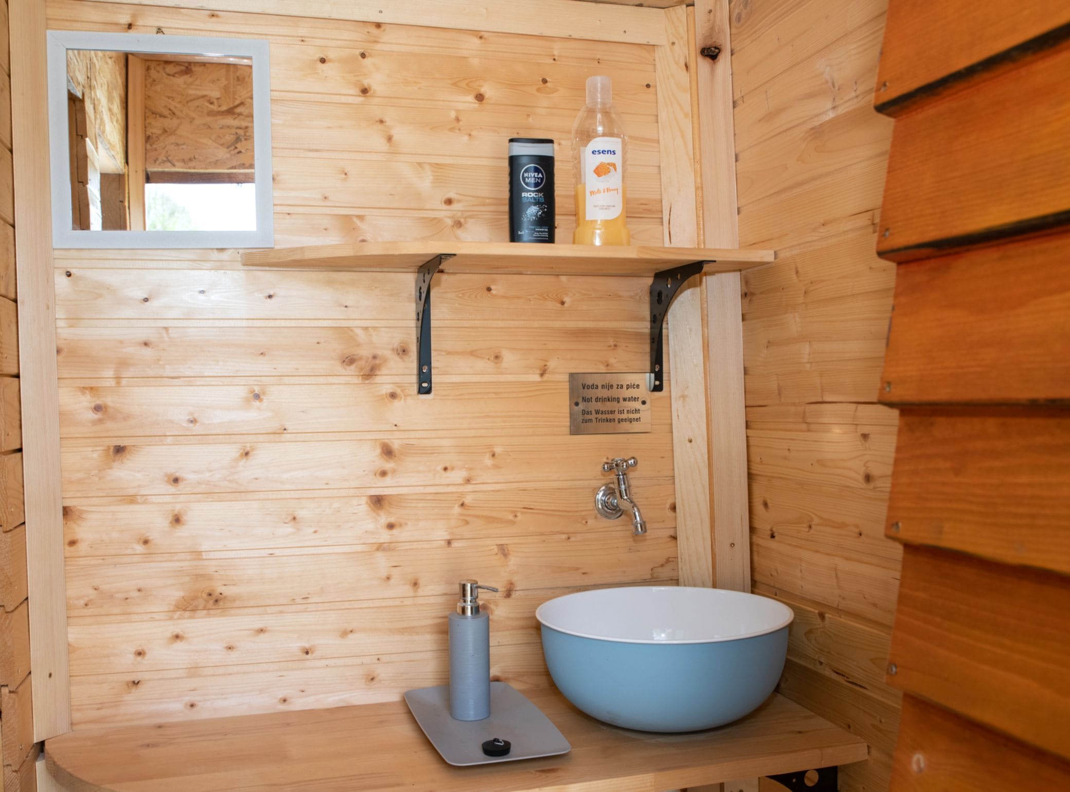 Baño de cabaña de madera con lavamanos, espejo, estante con artículos de aseo y señalización de agua.
