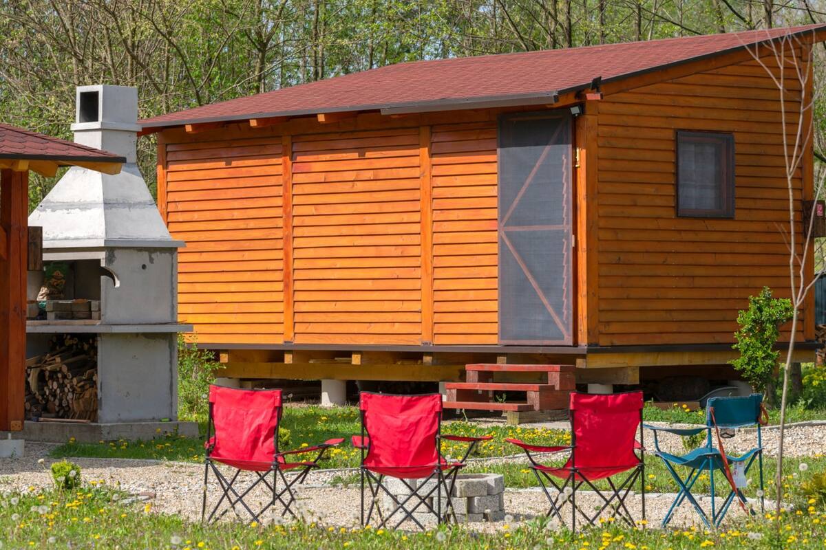 Eine Holzhütte mit Schornstein, kleinem Fenster und einer Tür. Vor der Hütte stehen rote und blaue Klappstühle.