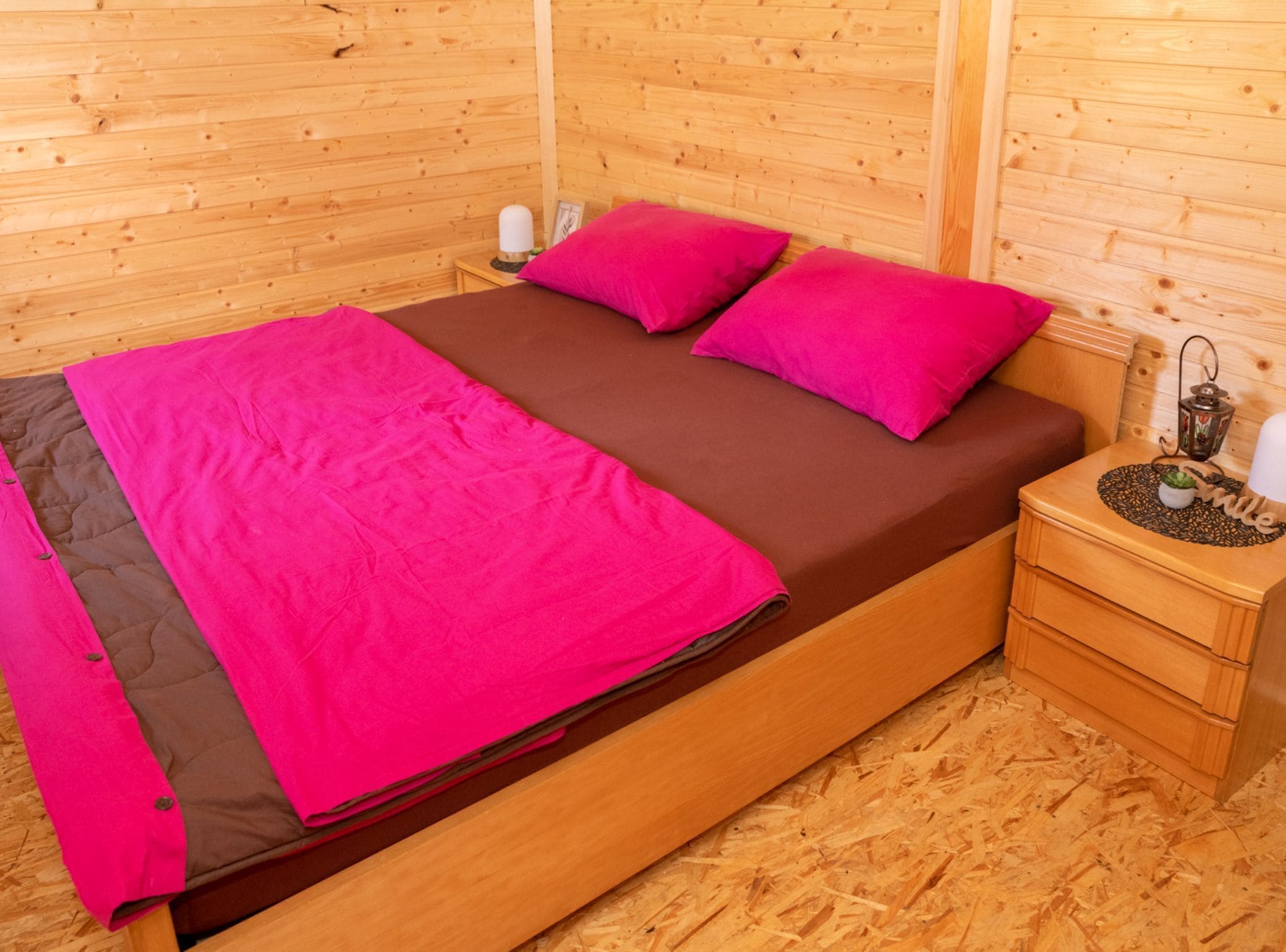 Ein holzgetäfeltes Zimmer im Blockhausstil mit einem Bett, rosa-brauner Bettwäsche, einem Nachttisch mit einer Laterne und einer kleinen Pflanze.