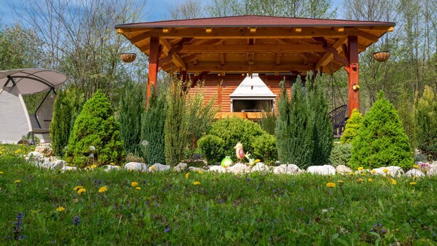 A wooden cabin with a covered patio, surrounded by greenery and trees.