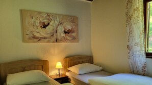 Quarto com duas camas, uma mesa de cabeceira com abajur, um quadro floral na parede e uma janela com cortinas.