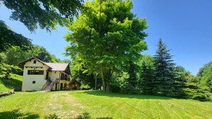 Uma casa de dois andares com uma varanda, cercada por um gramado e árvores.