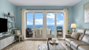 Een moderne woonkamer met een bank, salontafel en tv-meubel. Door grote ramen is een balkon met uitzicht op de stad en zee te zien.