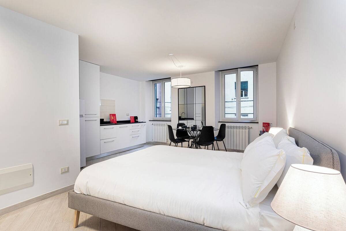 A modern bedroom with a large bed, a dining table with chairs, a kitchen area, and a view of the city through the windows.