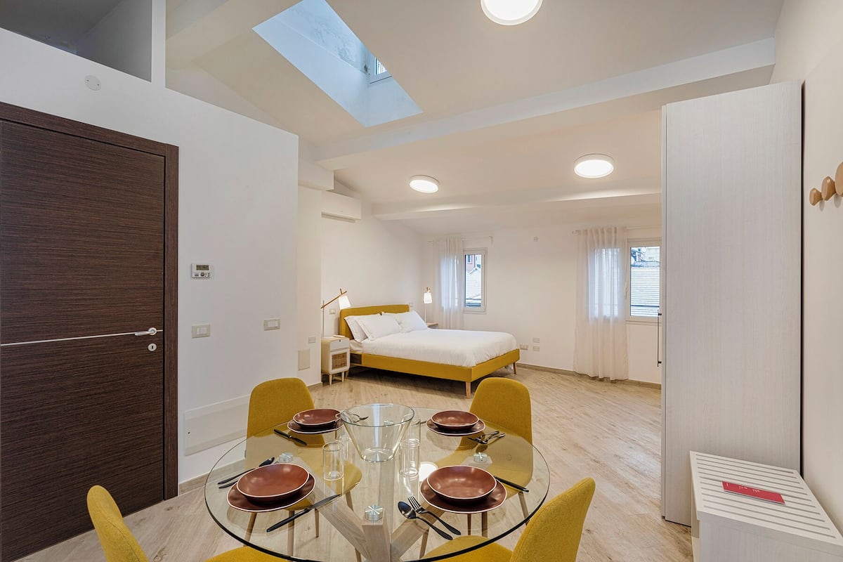 A modern bedroom with a bed, a skylight, and a dining area with a glass table and chairs.