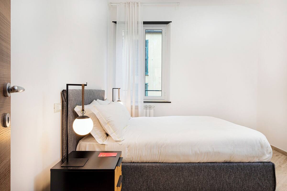 A bedroom with a bed, a bedside table with a lamp, a window with curtains, and a view of buildings outside.