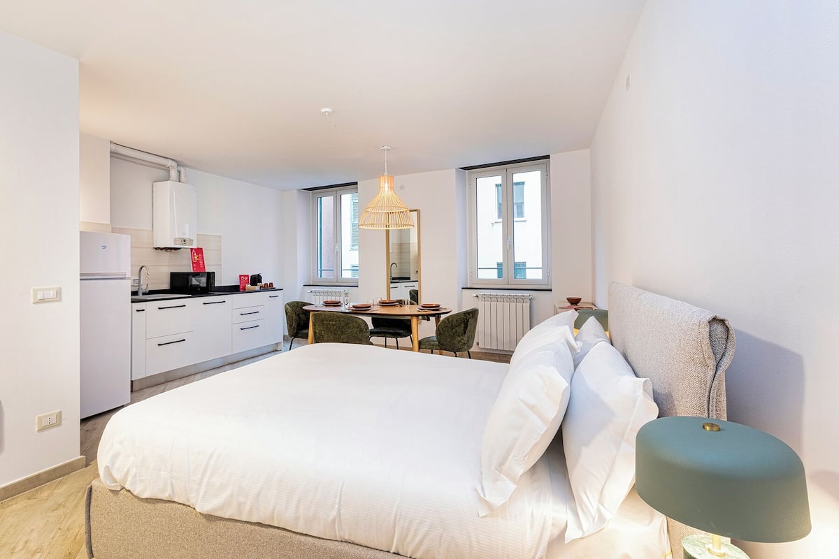 A modern bedroom with a large bed, a bedside lamp, and a view of the kitchen and dining area.