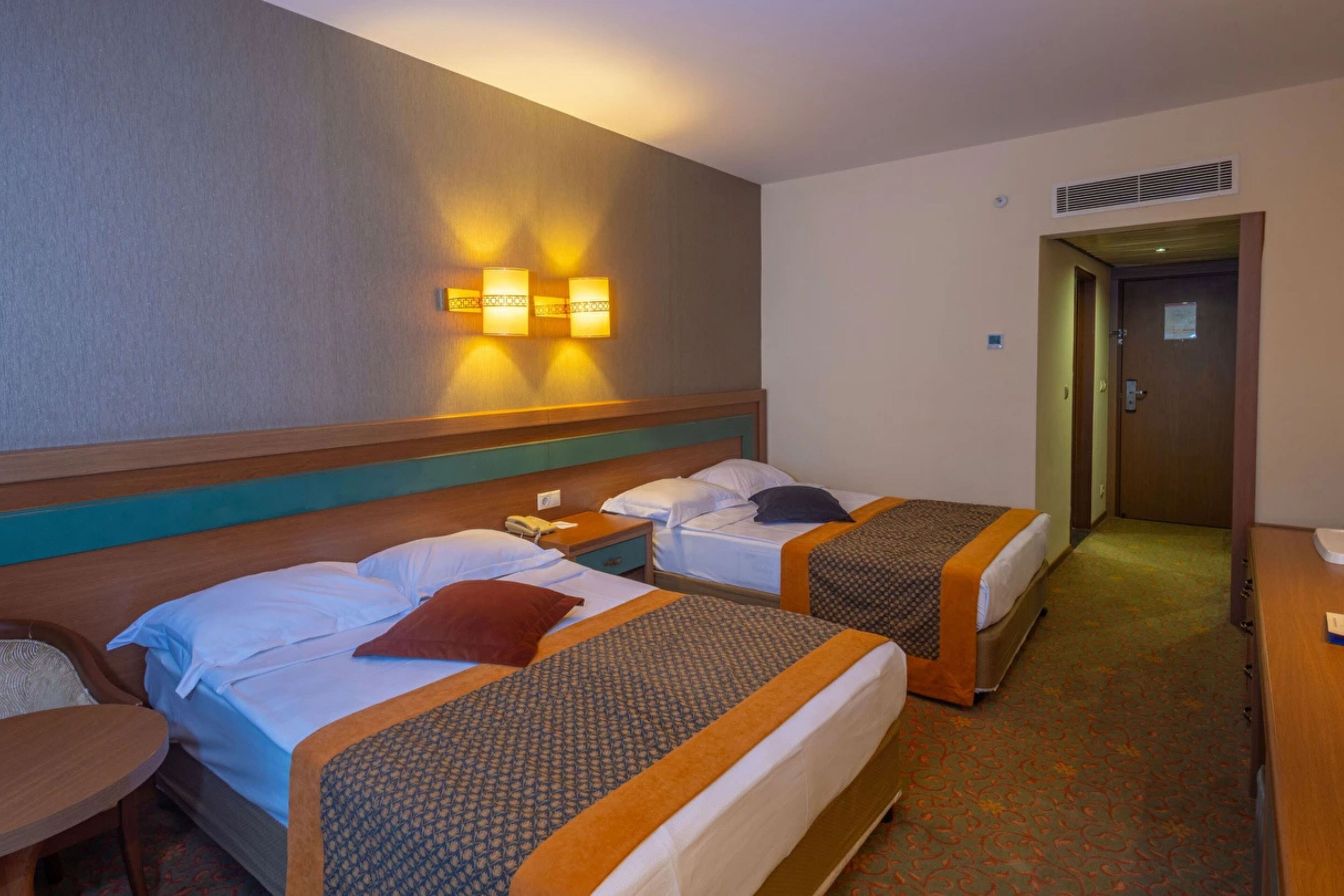 A hotel room with two beds, a desk, a chair, and a wall-mounted light fixture.