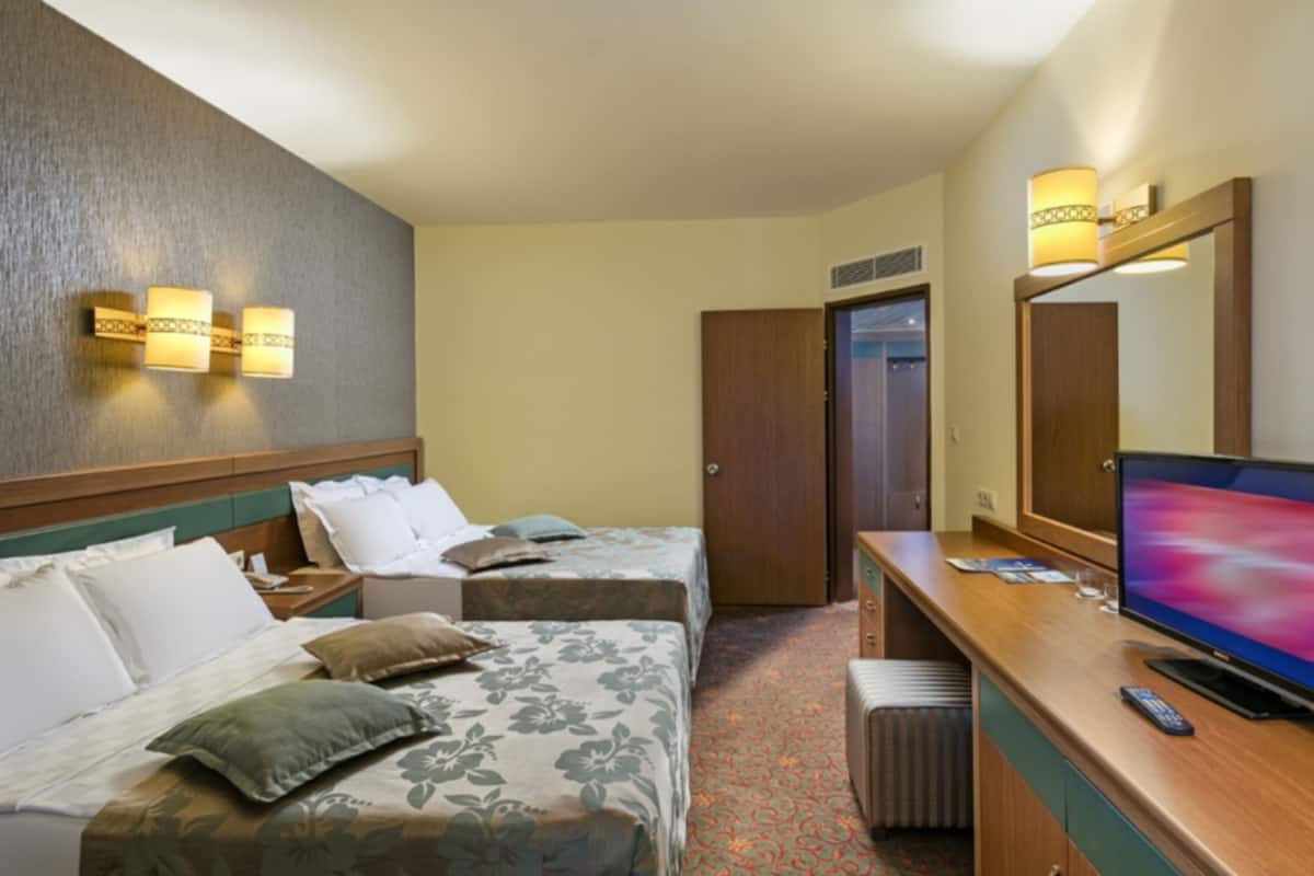 A hotel room with two beds, a desk with a television, and wall-mounted lighting.