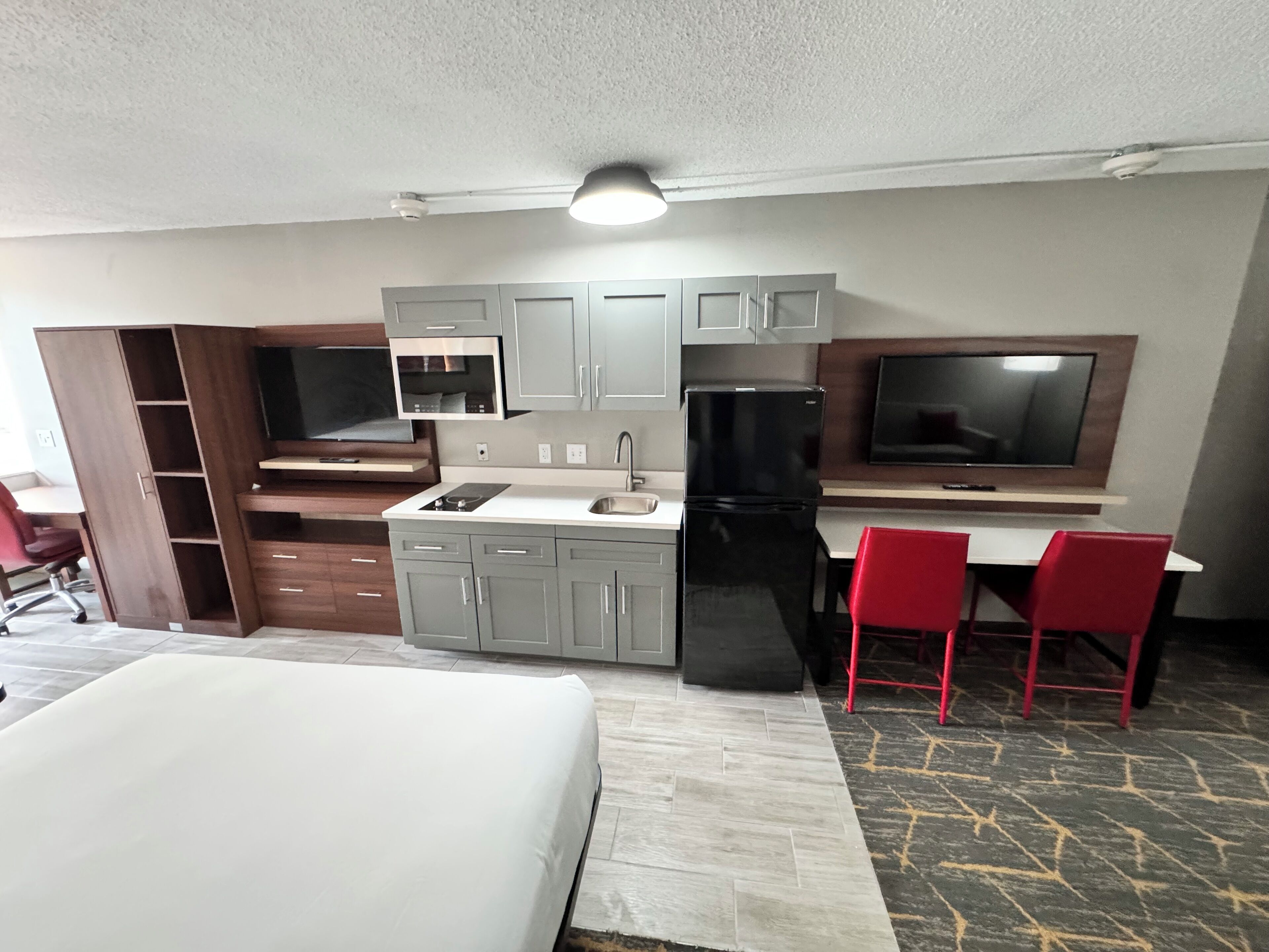 Cocina compacta con armarios grises, un refrigerador negro, microondas y televisor integrado. Hay dos sillas rojas y una cama con ropa de cama blanca.