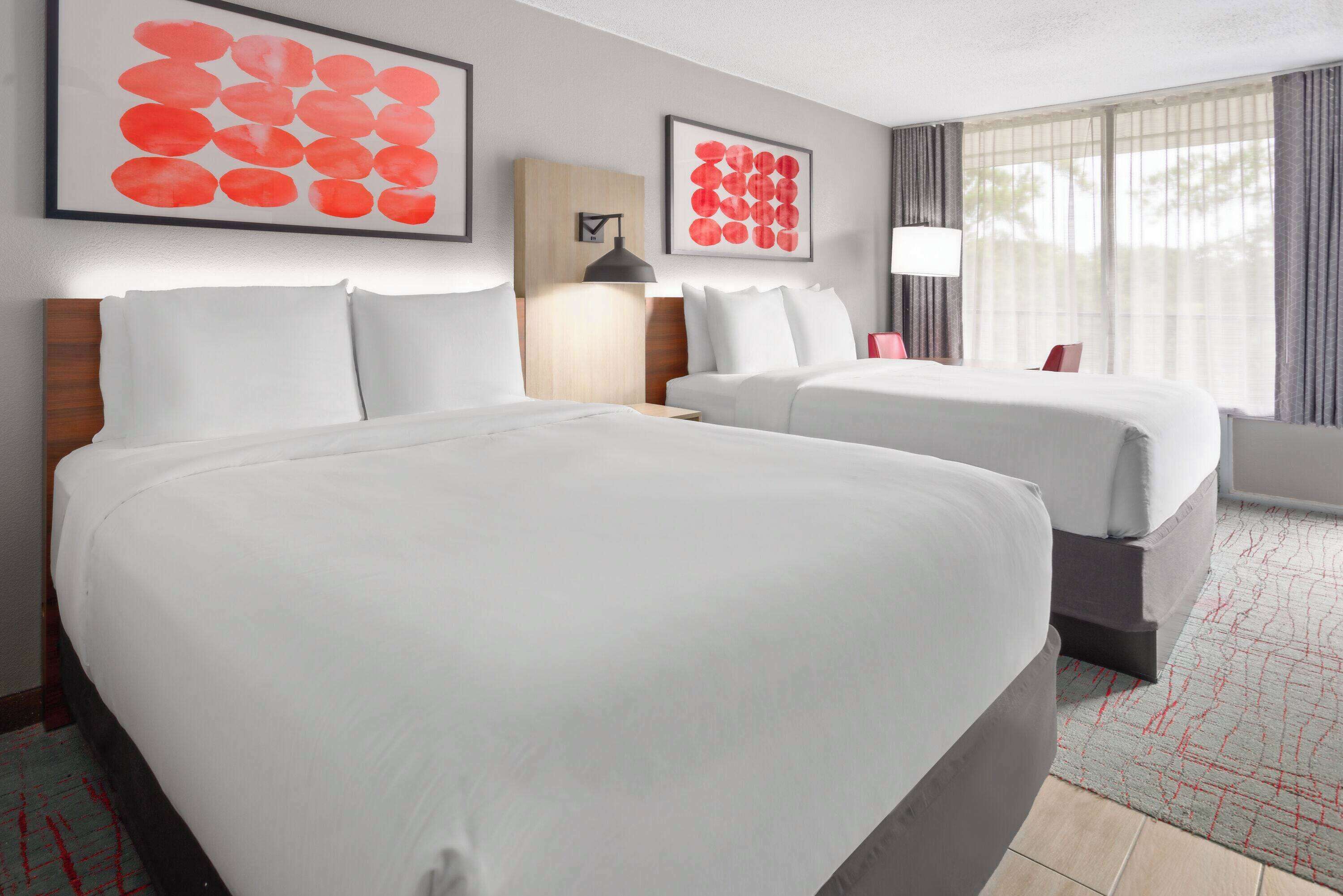Habitación de hotel con dos camas, un ventanal con cortinas y dos cuadros abstractos rojos en la pared.
