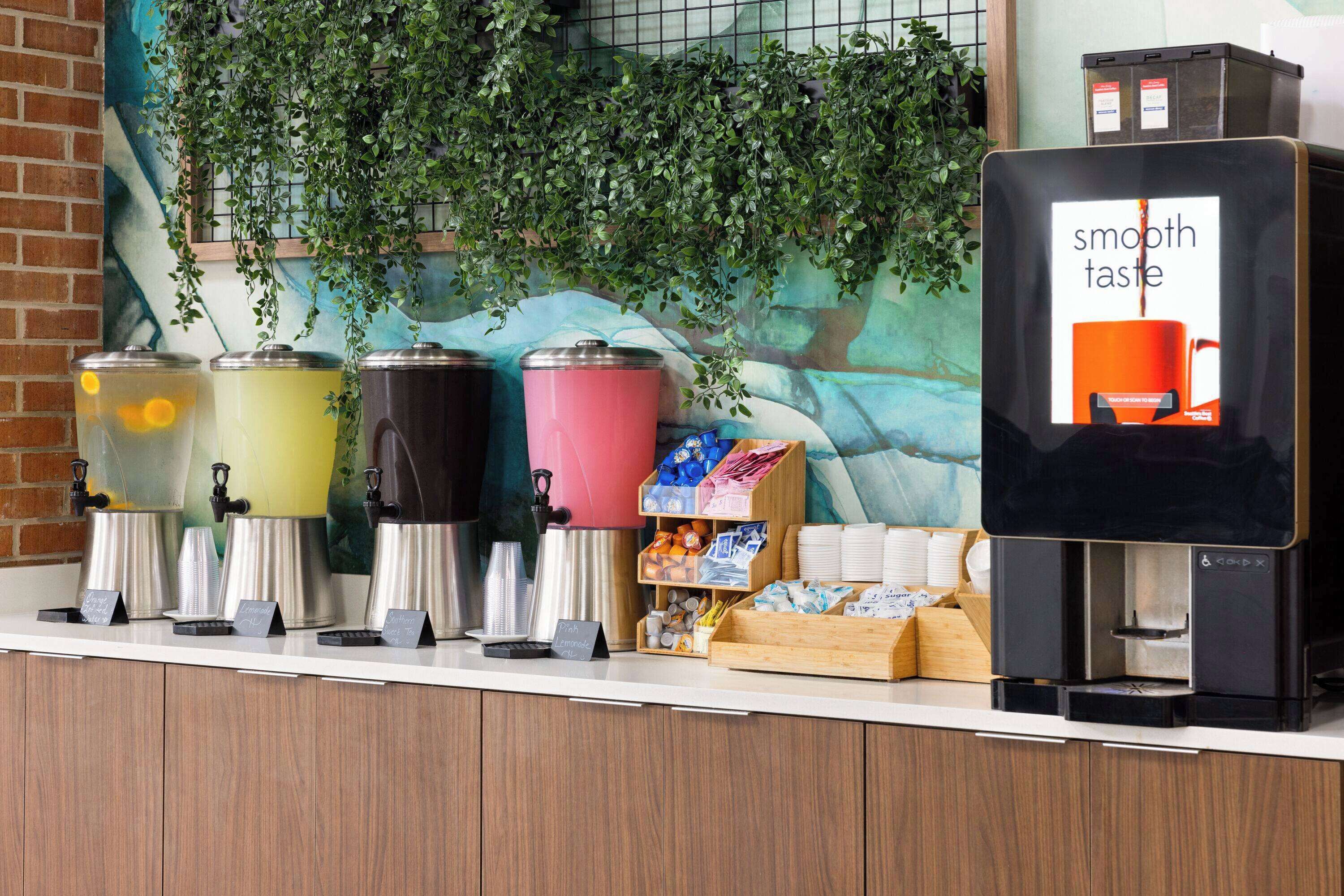 A beverage station with multiple dispensers and a coffee machine, surrounded by a counter with various items and a mural in the background.