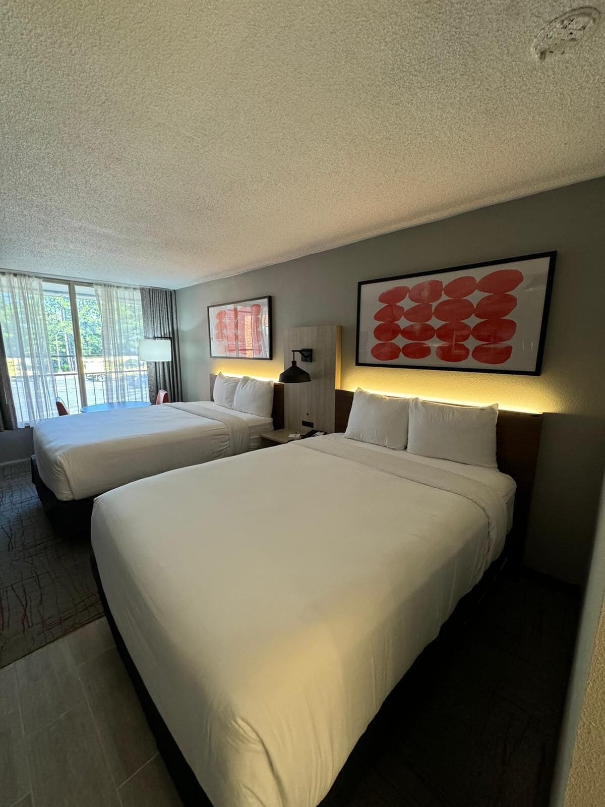 A hotel room with two beds, a large window with curtains, a wall-mounted lamp, and a framed artwork.