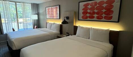 A hotel room with two beds, a large window with curtains, a wall-mounted lamp, and a framed artwork.