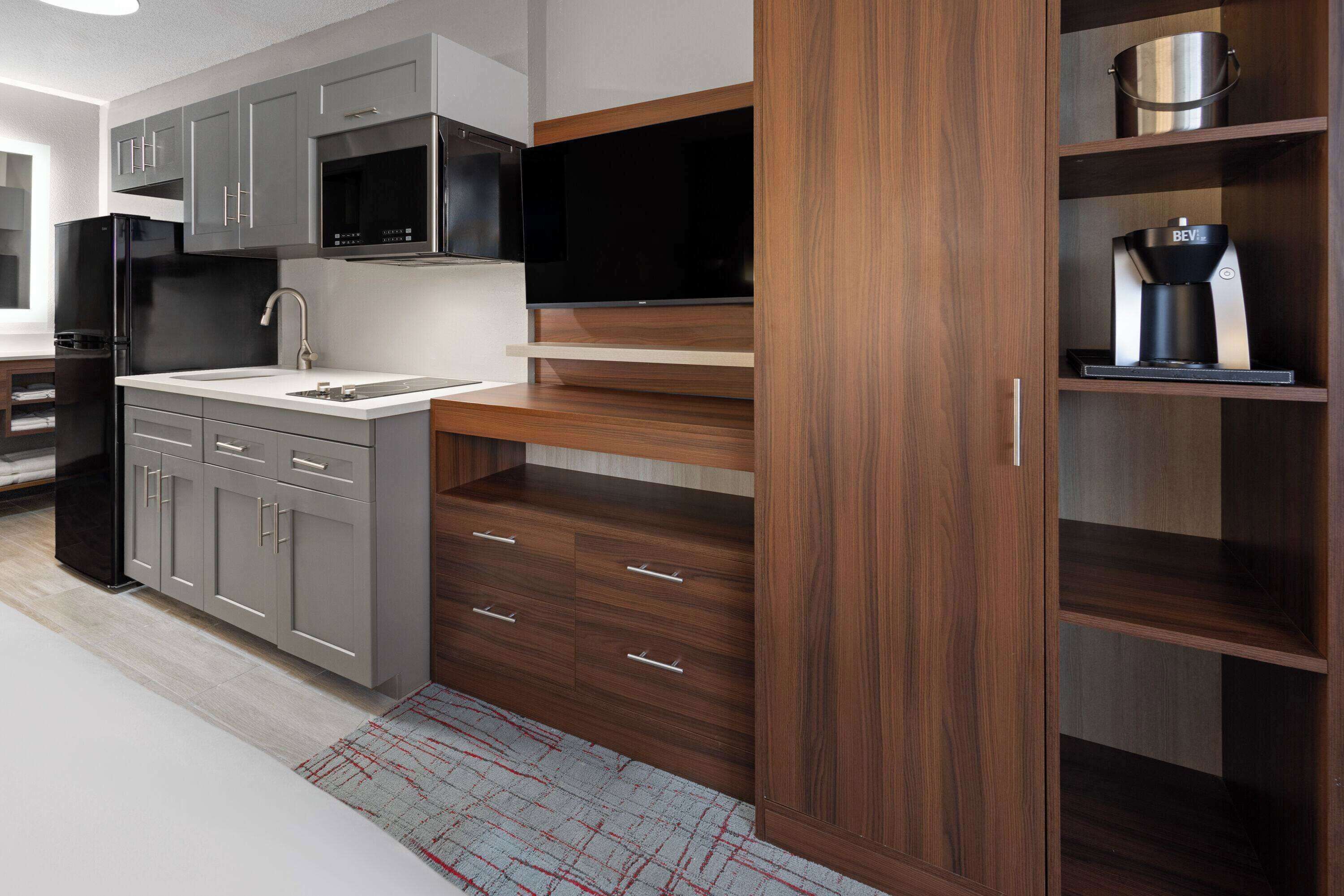 A modern kitchen with grey cabinets, a black refrigerator, a microwave, a built-in oven, and a coffee maker on a wooden shelf.