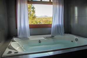 A modern bathroom with a large bathtub, a window with curtains, and a view of a natural landscape. - Pousada Canto Verde Cipó (Jaboticatubas)