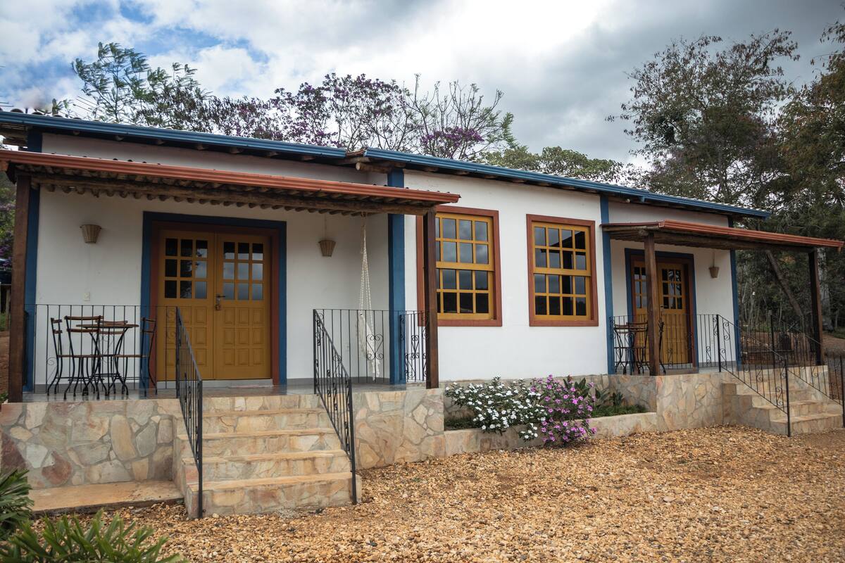 Una casa de dos pisos con base de piedra, puertas de madera y un porche cubierto con mobiliario exterior.