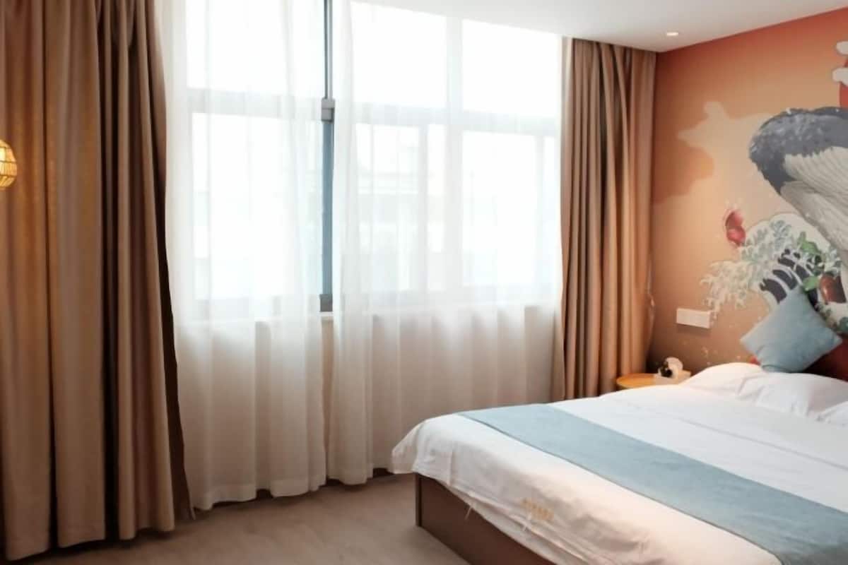 Quarto de hotel com uma cama grande, janela com cortinas finas, um mural na parede e uma mesa de cabeceira com abajur.