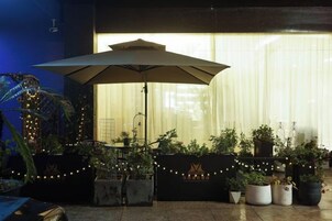 Un área de estar al aire libre con una sombrilla grande, plantas en macetas y luces colgantes.