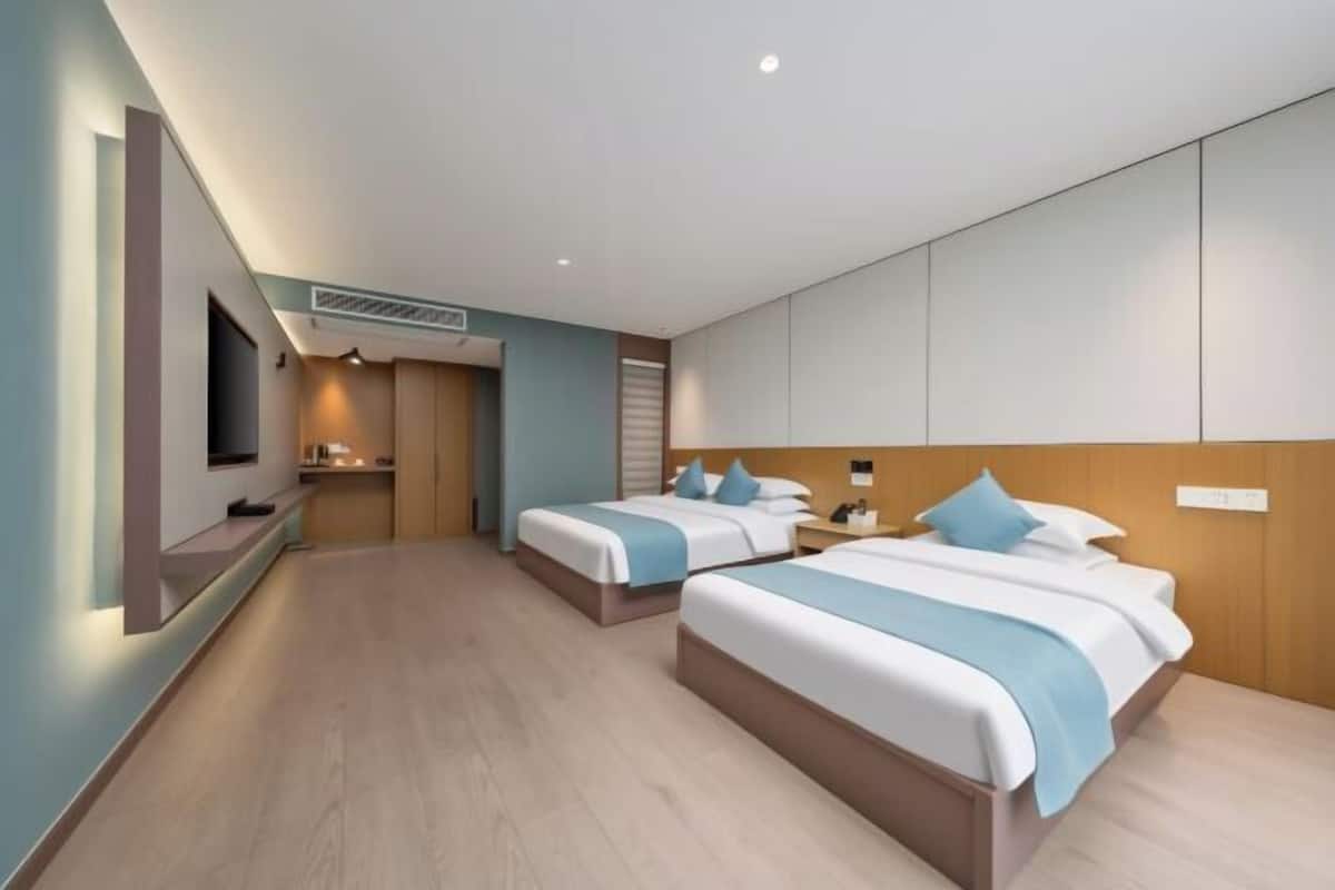 Quarto de hotel com duas camas, televisão, pequena cozinha e cabeceira de madeira.