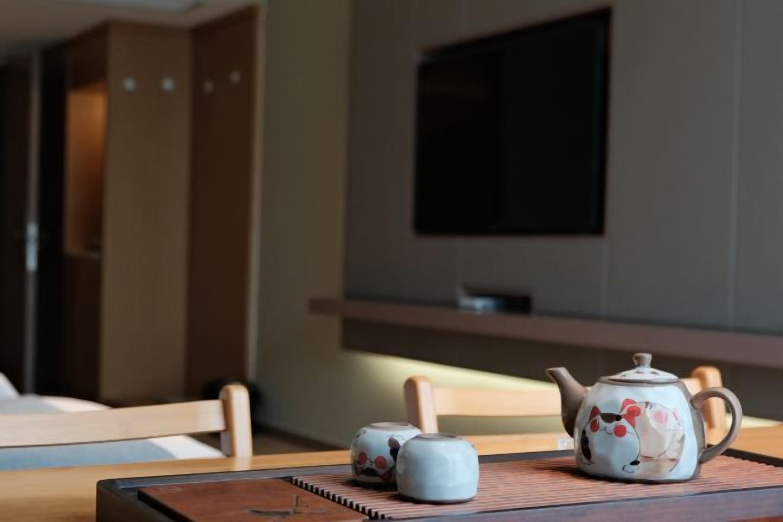 Una tetera y dos tazas sobre una mesa de madera, con un televisor al fondo.