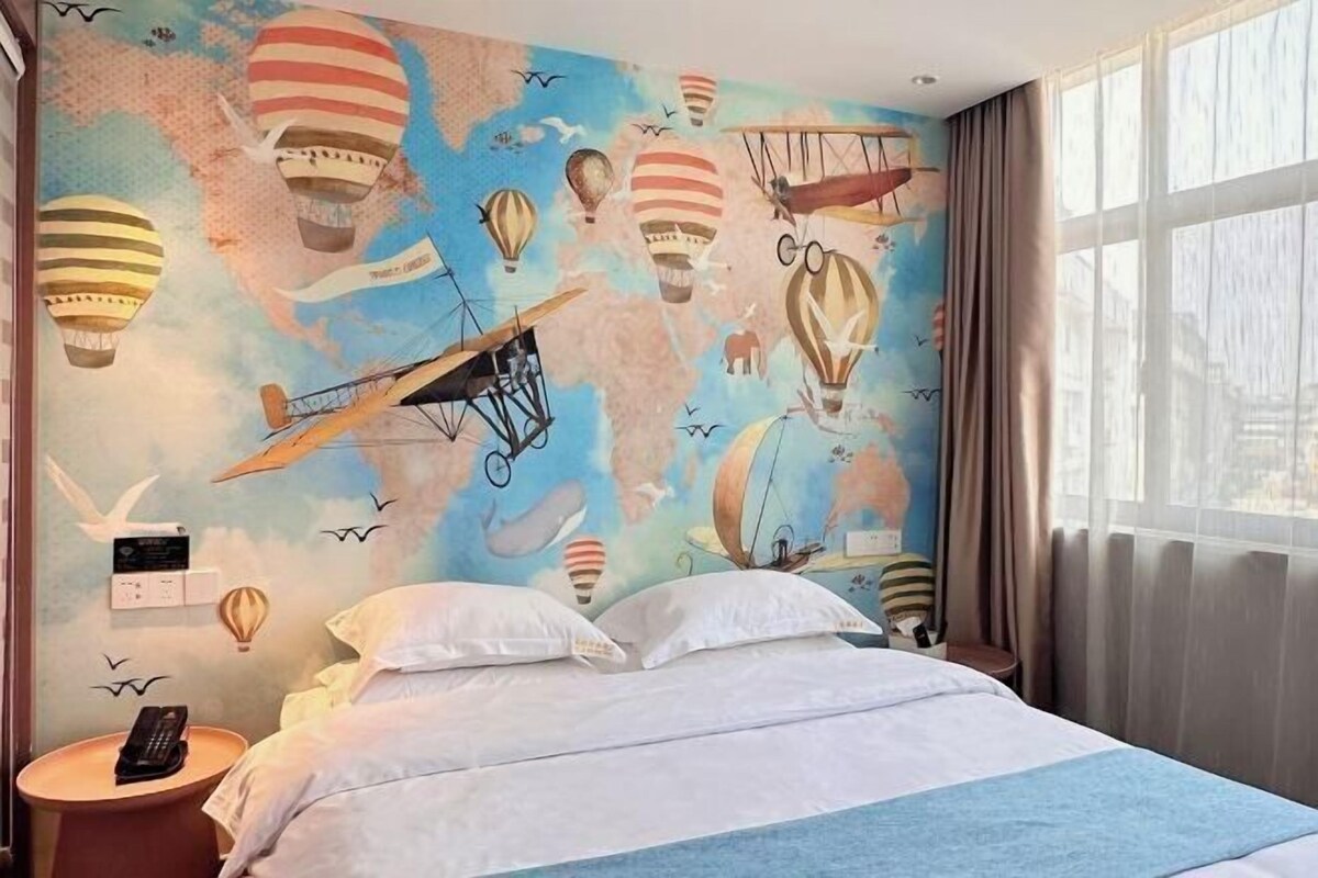 A hotel room with a bed, a bedside table with a phone, and a mural of hot air balloons and airplanes on the wall.
