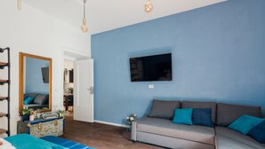 Et værelse med blå væg, en grå sofa med turkise puder, et fladskærms-tv monteret over sofaen, trægulv og et stort spejl med blomstret kommode.