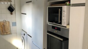 A compact kitchen with a washing machine, microwave, and oven built into white cabinetry. - Maison Fond de Jardin Située à 100 m de la Plage. 2 Chambres Pour 7 Personnes (Mimizan)