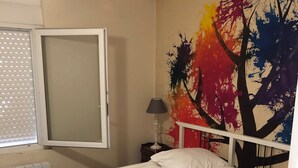 A single-bed room with a white mattress, a white headboard, a window with blinds, a radiator, a nightstand with a lamp, and a colorful abstract painting on the wall. - Maison Fond de Jardin Située à 100 m de la Plage. 2 Chambres Pour 7 Personnes (Mimizan)