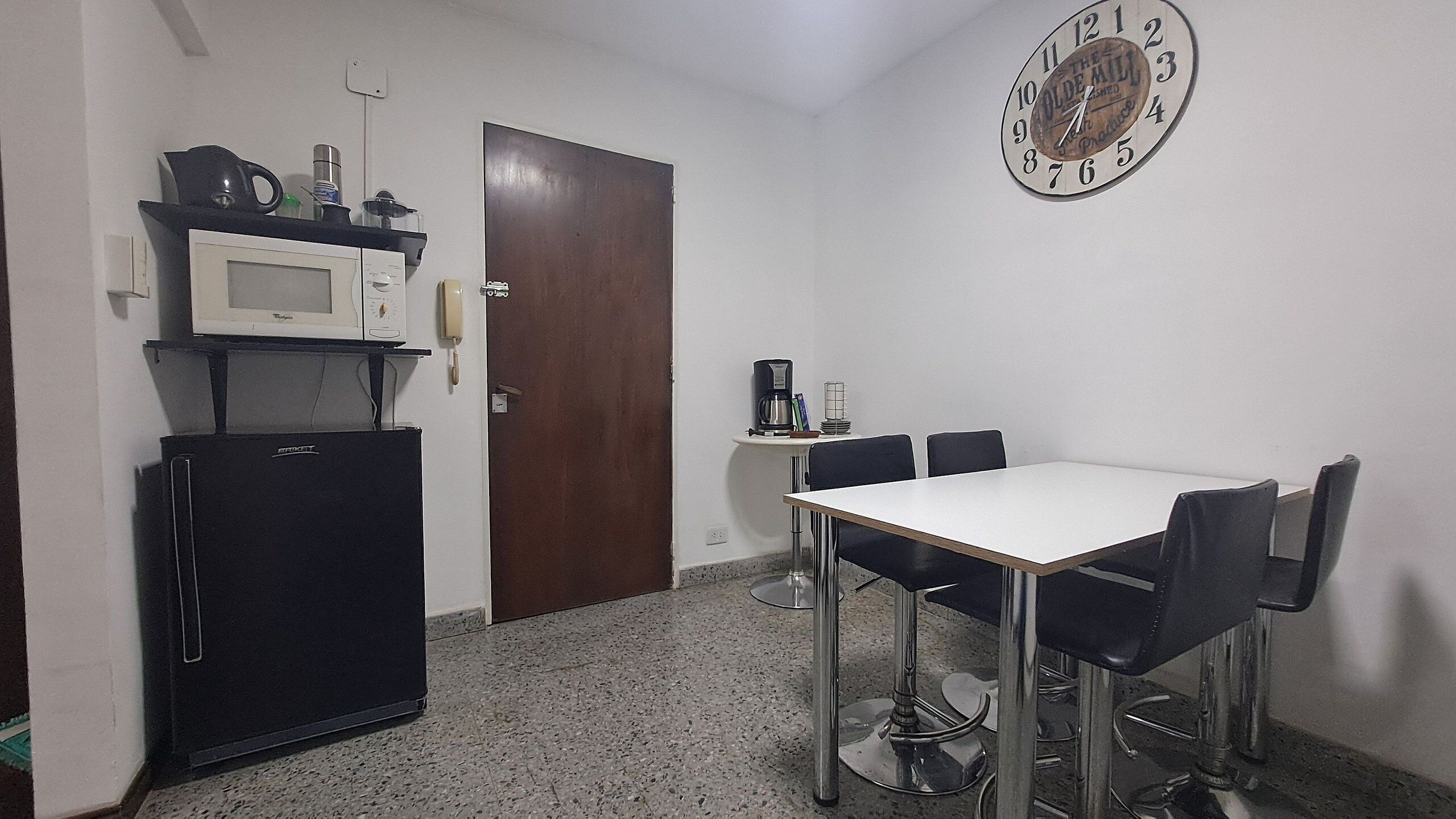 Een kleine kitchenette met een magnetron, een koelkast en een koffiezetapparaat. Er is een eettafel met vier stoelen en een klok aan de muur.