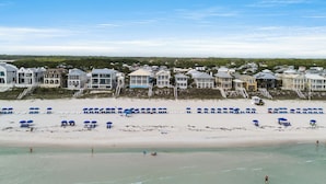 Ein Strand mit blauen Liegestühlen und Sonnenschirmen, ein sandiger Uferstreifen und Häuser im Hintergrund.