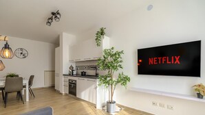 Un salon moderne doté d’un téléviseur à écran plat affichant le logo « NETFLIX » sur le mur, d’un coin repas avec des chaises et une table, et d’une cuisine avec des meubles blancs et un plan de travail noir.