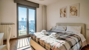 Une chambre avec un grand lit, une vue sur la ville par une porte coulissante, un radiateur et deux œuvres d’art abstraites accrochées au mur.
