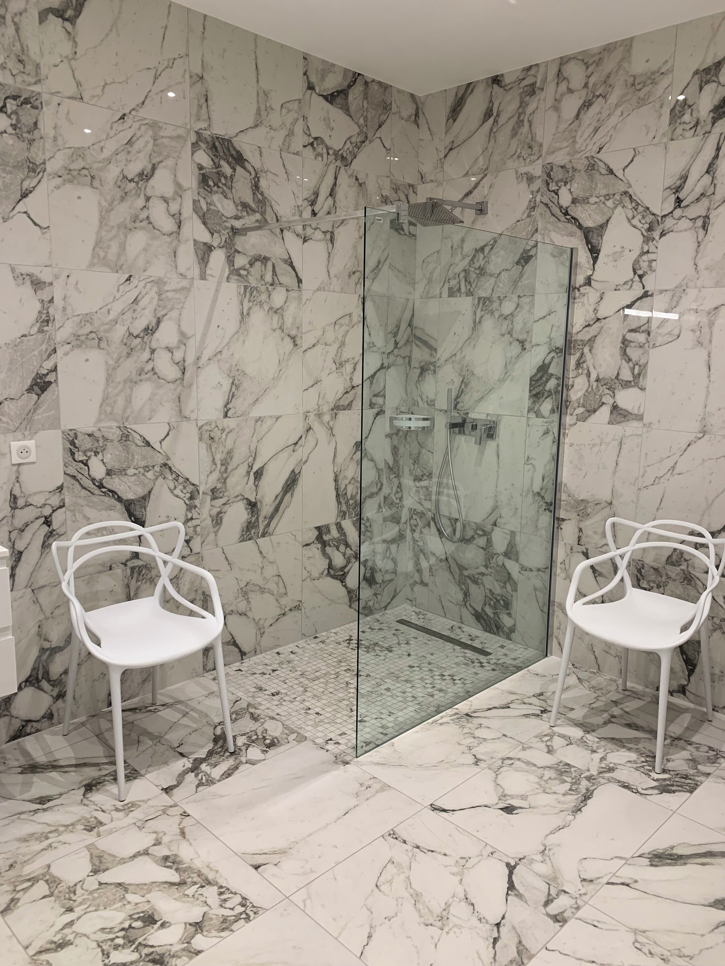 Ein modernes Badezimmer mit Marmorwänden und -boden, einer Duschkabine aus Glas sowie zwei weißen Stühlen.