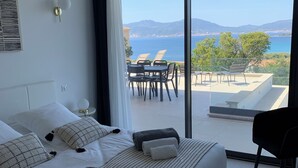 Una habitación de hotel moderna con una cama grande, un balcón con vistas al mar y a las montañas, y un área de comedor con sillas y una mesa.