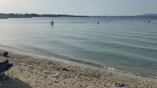 A beach with clear water, a sandy shore, and a distant city skyline. - Bedesten Pansiyon (Cesme)