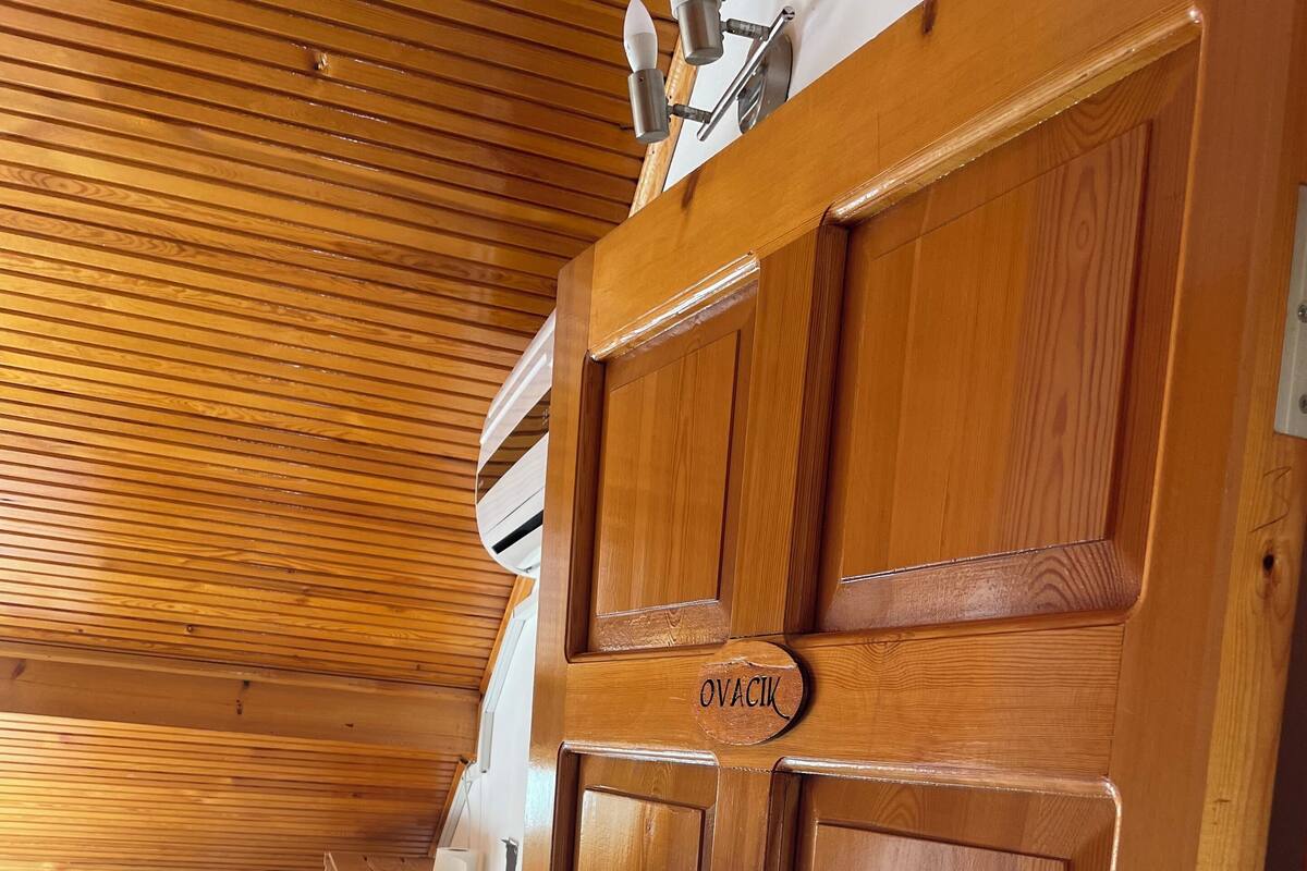 A wooden door with the name "OVACIK" on it, a wooden ceiling, a radiator, a small table with a white tablecloth, and a white appliance in the background.