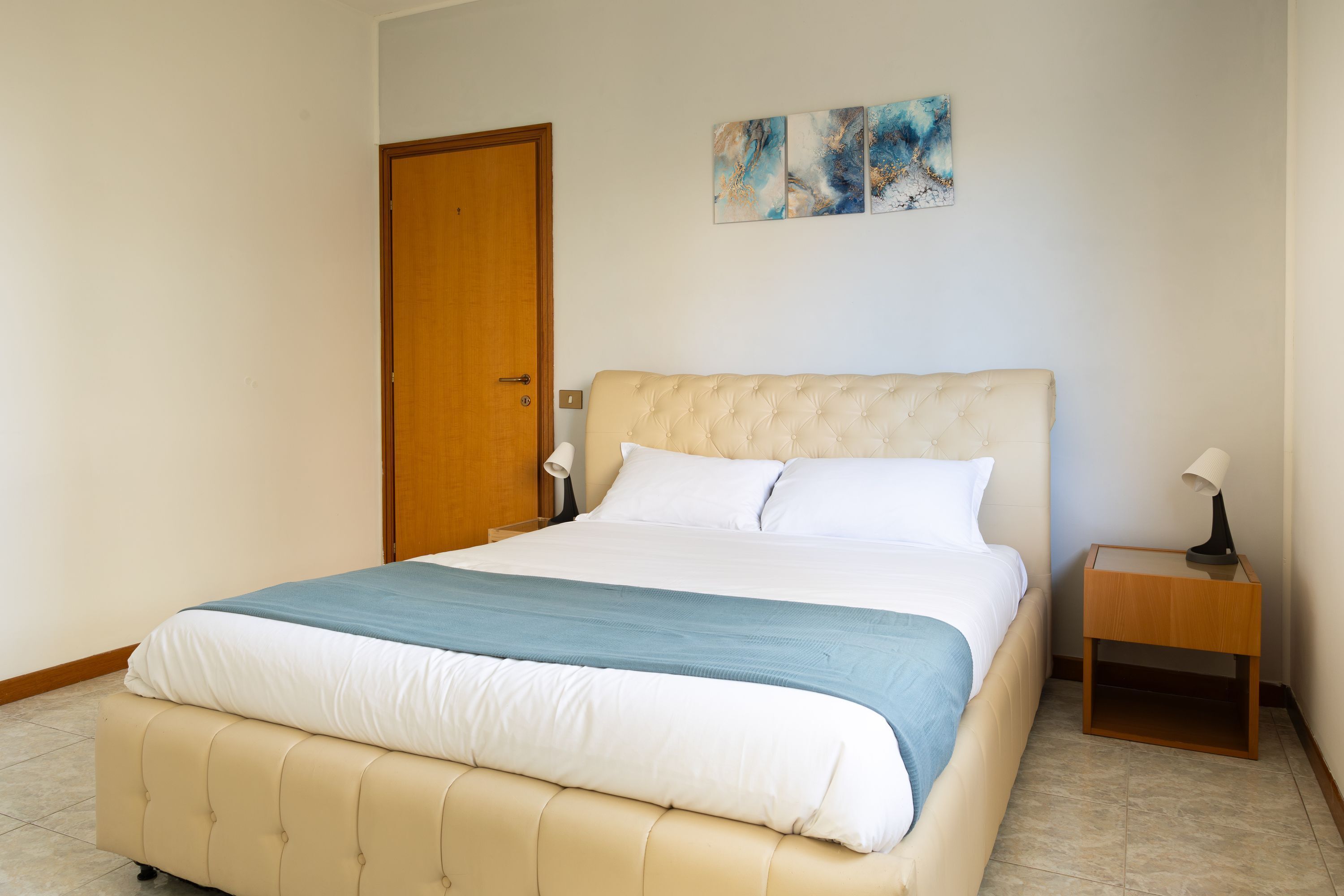 A bedroom with a large bed, a wooden door, a nightstand with a lamp, and abstract wall art.