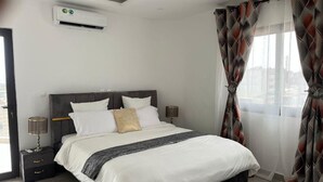 Une chambre d’hôtel moderne avec un grand lit, une table de chevet avec une lampe et une vue sur la ville à travers des rideaux transparents.