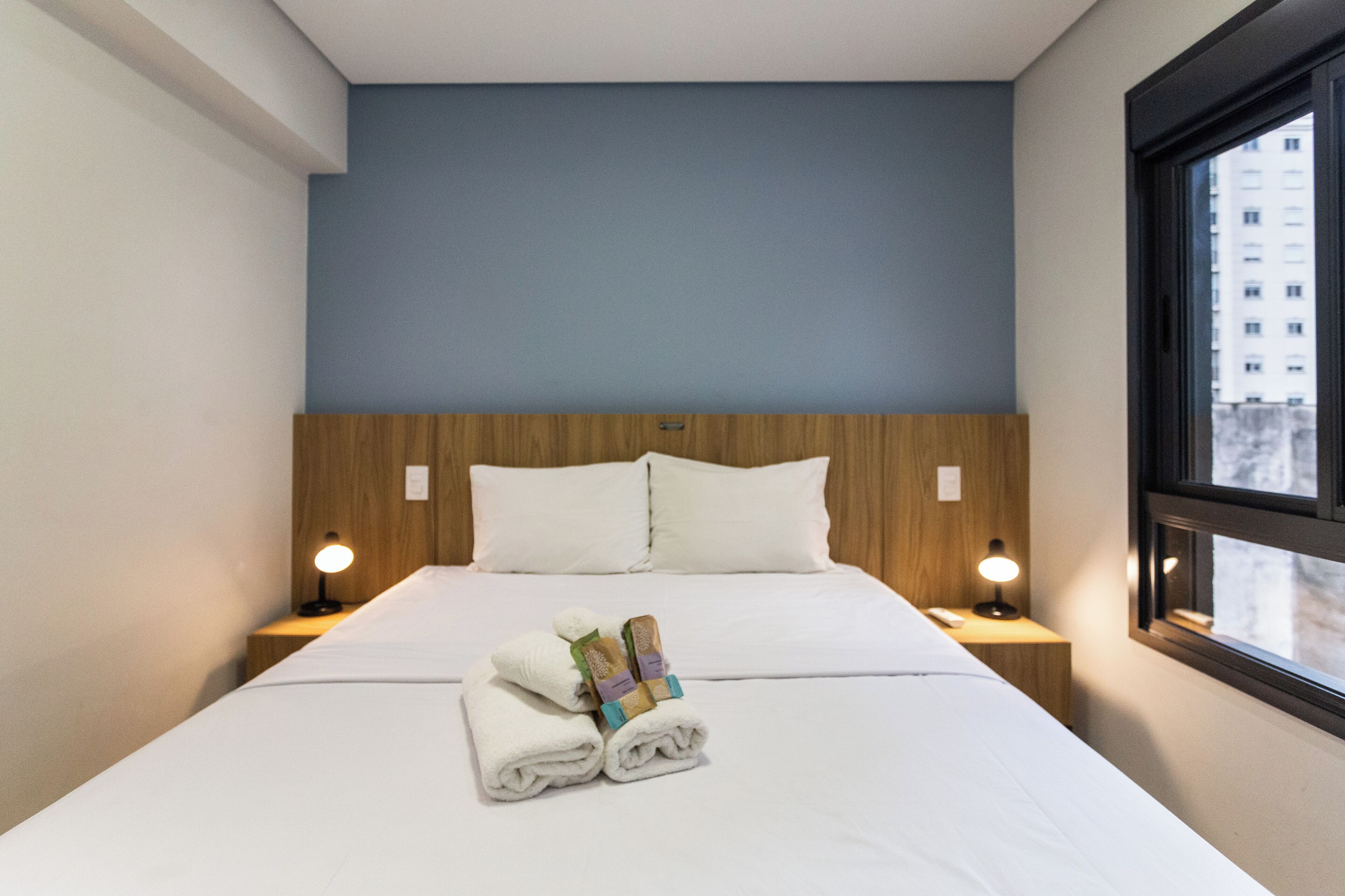 Una camera d'albergo con un letto rifatto, due abat-jour e una finestra con vista sugli edifici.