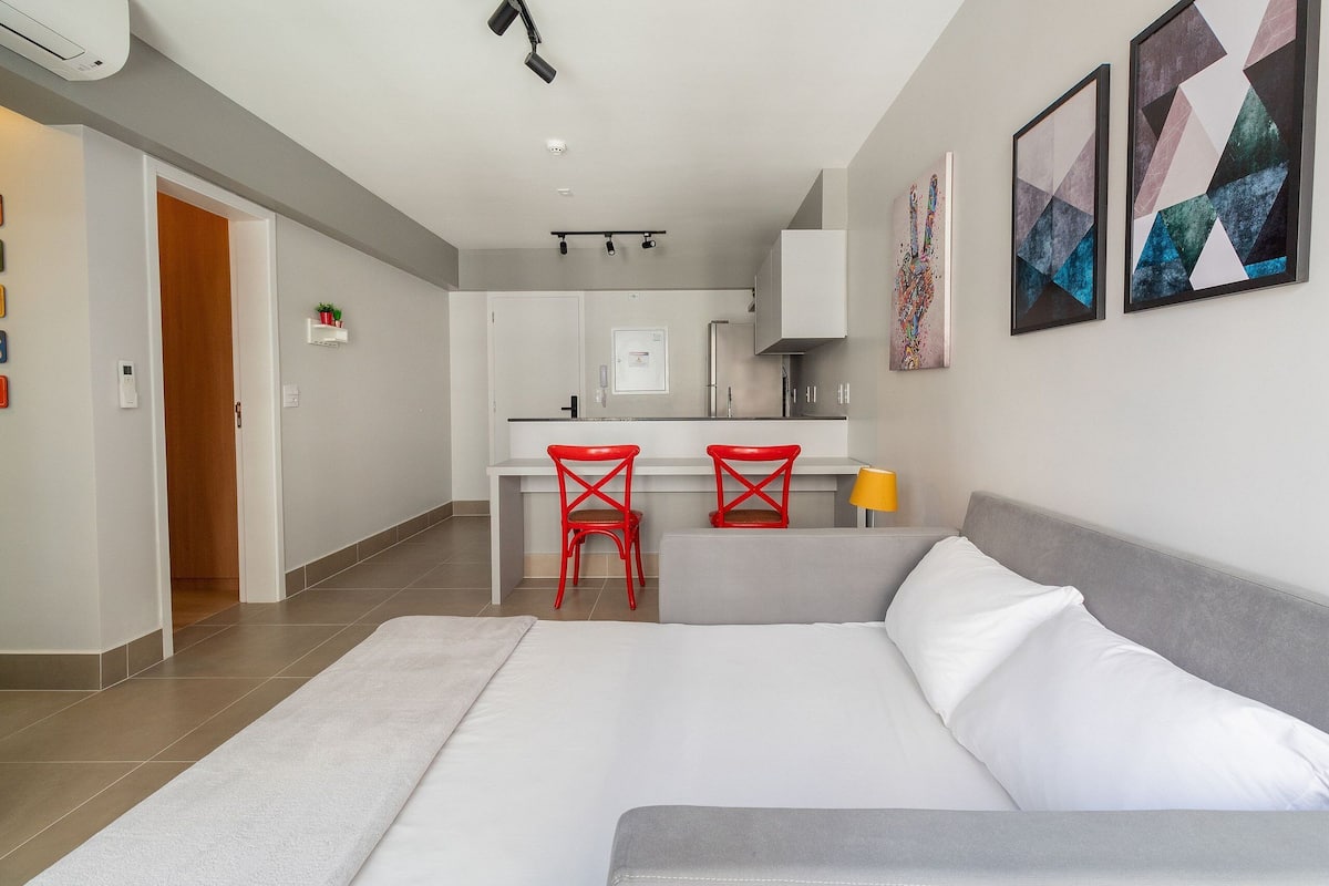 Moderni hotellihuone, jossa on sänky, työpöytä, kaksi punaista tuolia ja taideteoksia seinillä.