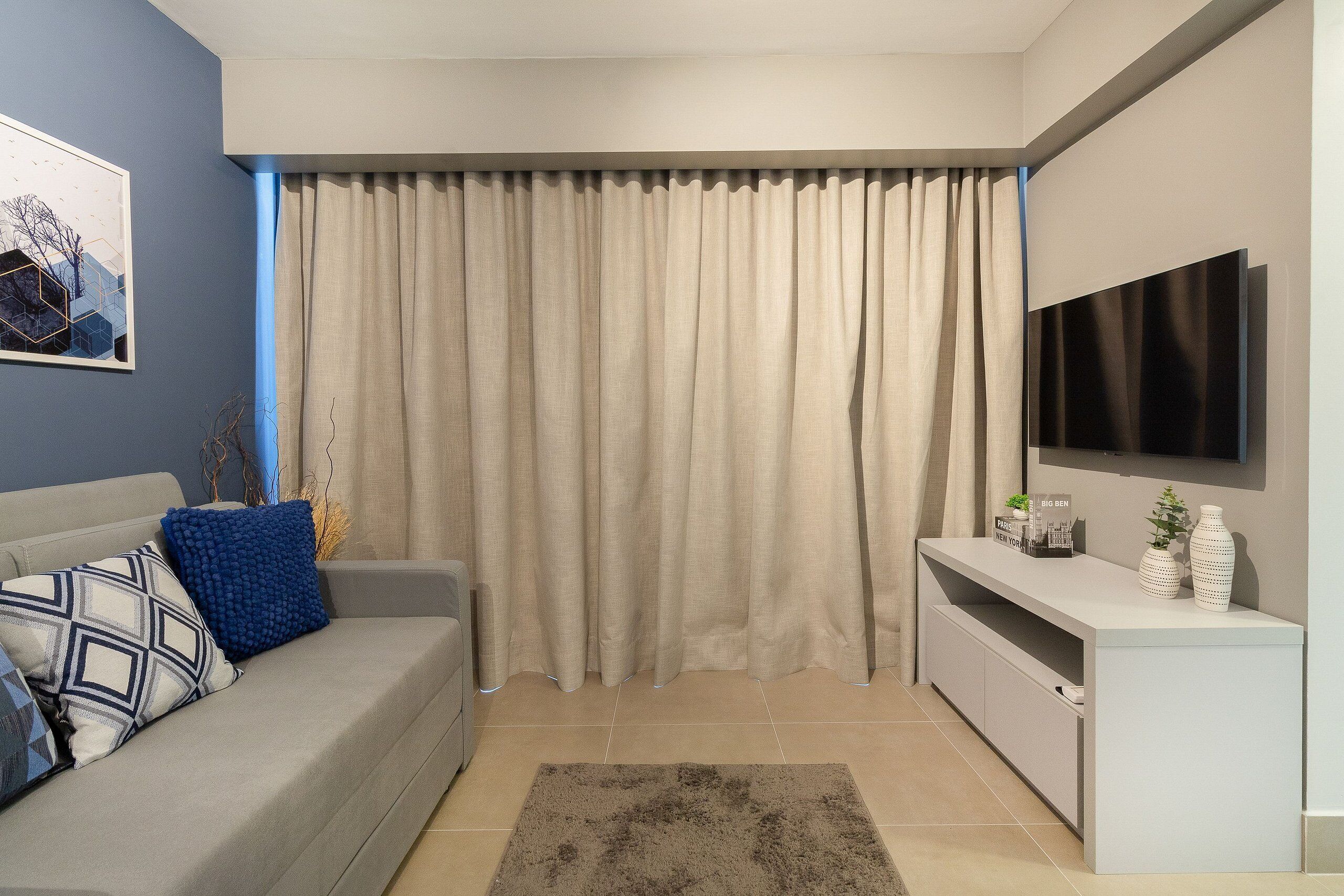 A modern living room with a grey sofa, a coffee table, a TV on a stand, and a large window with beige curtains.